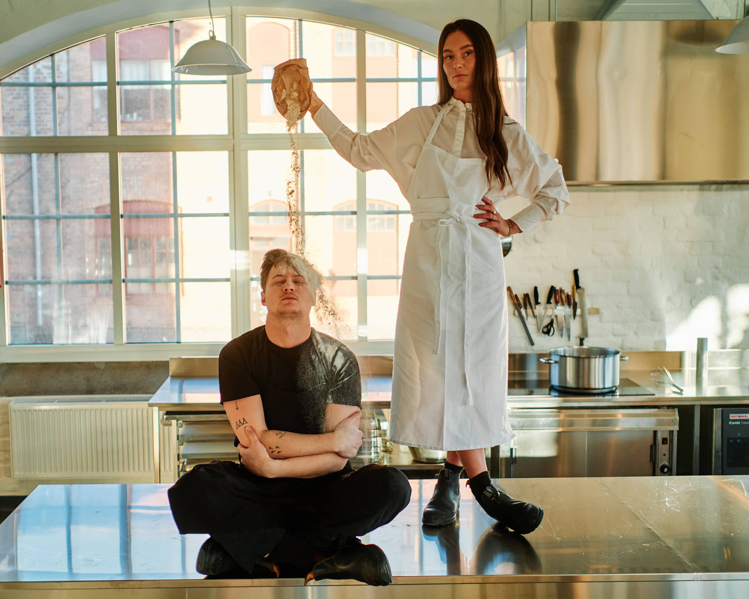 Evelina standing on kitchen counter at Salt Studio Gävle pouring flour, Rasmus sitting cross-legged below